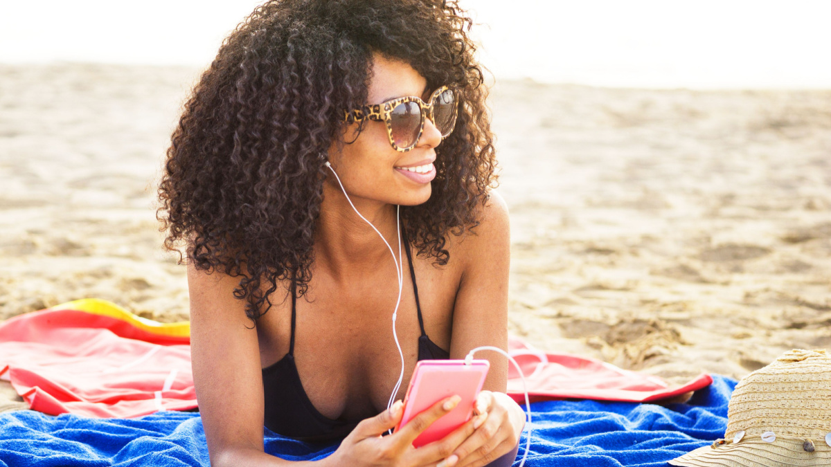 african origin girl relaxing on beach. afro hair woman wearing sunglasses lying on beach using phone listening to trendy music with ear phones.