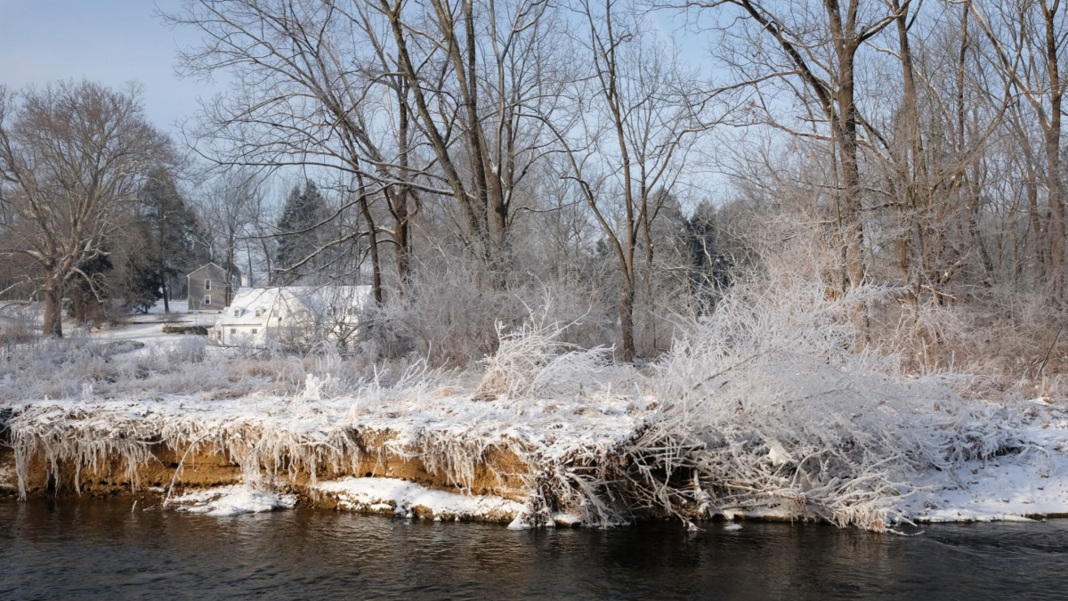 White house in sunlight in distance with snow covered grass and tress on the river bank in front