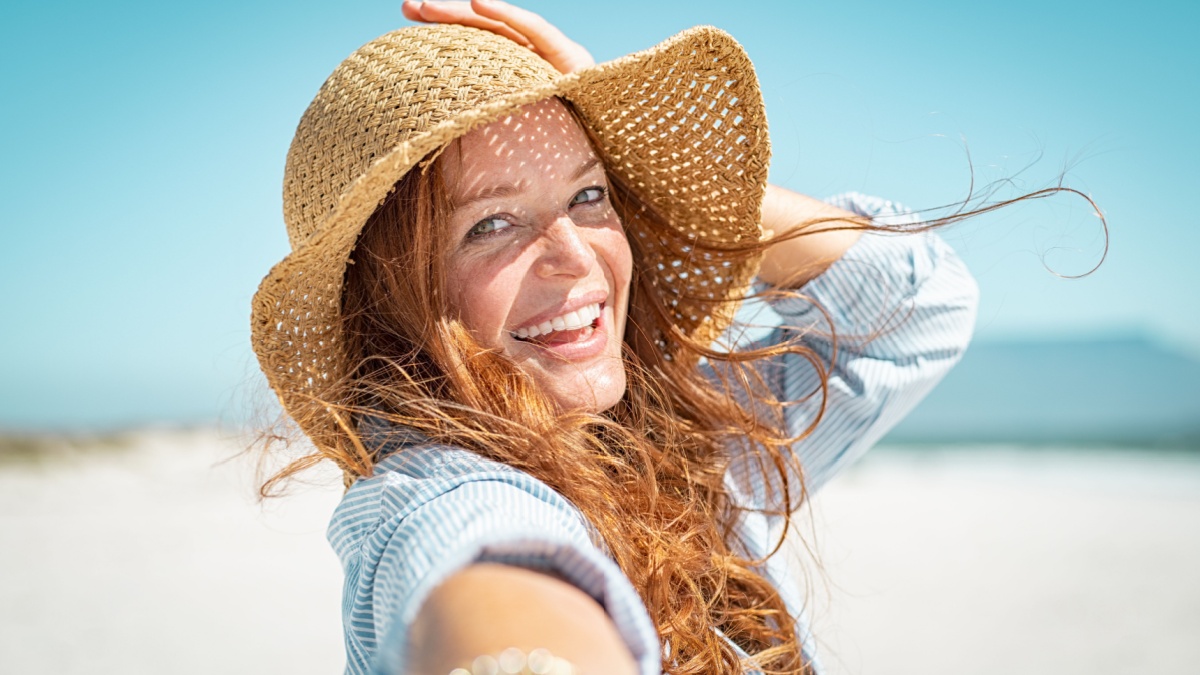 Portrait of beautiful mature woman in casual wearing straw hat at seaside. Cheerful young woman smiling at beach during summer vacation. Happy girl with red hair and freckles enjoying the sun.