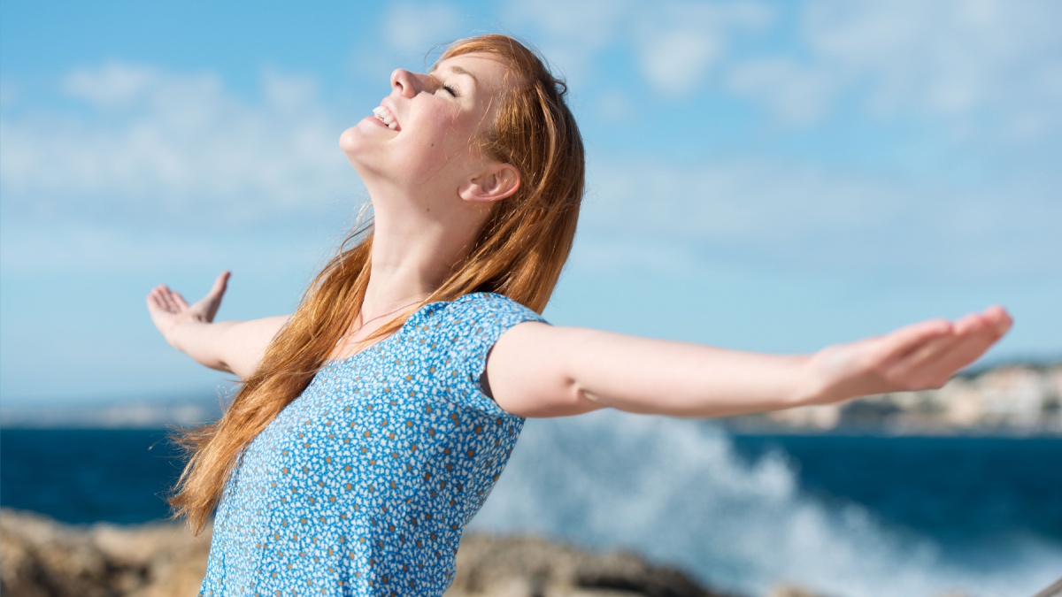 beautiful woman celebrating the sunshine standing with her arms outspread and eyes closed in bliss in front of breaking ocean waves.