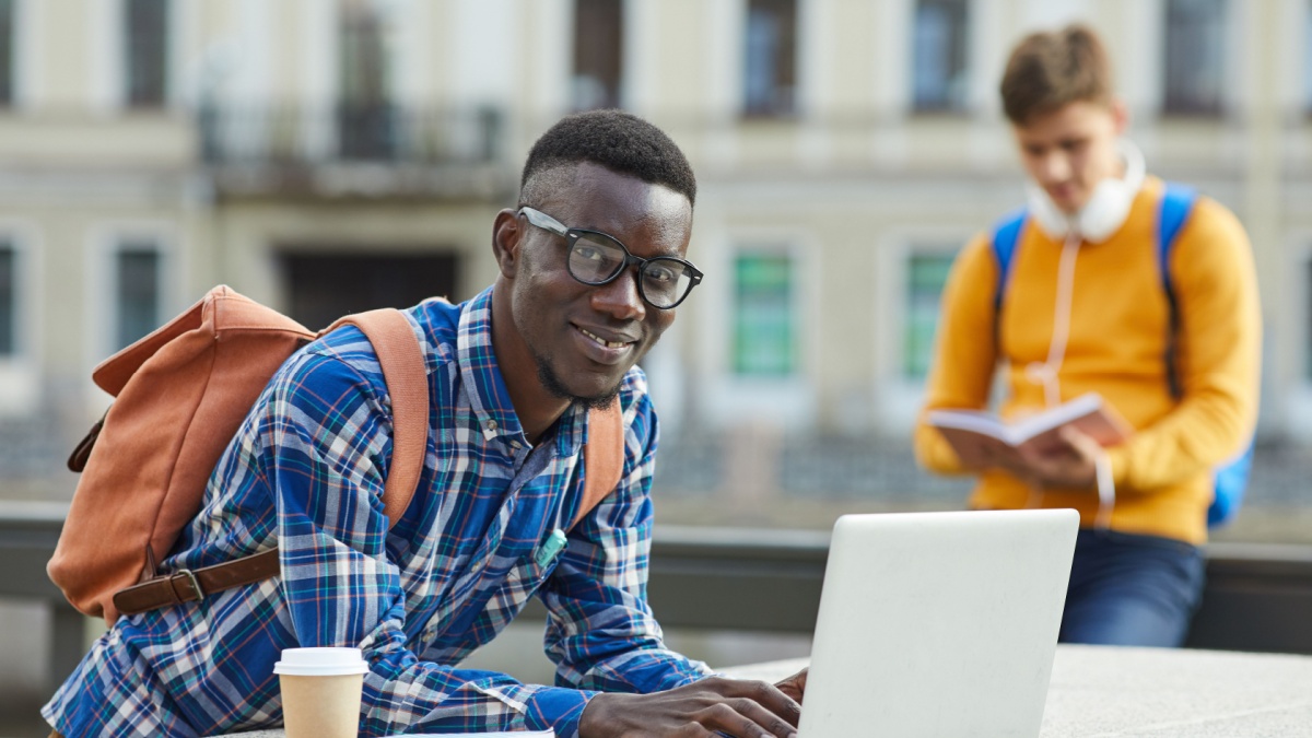 Portrait of contemporary African student using laptop outdoors in college campus.