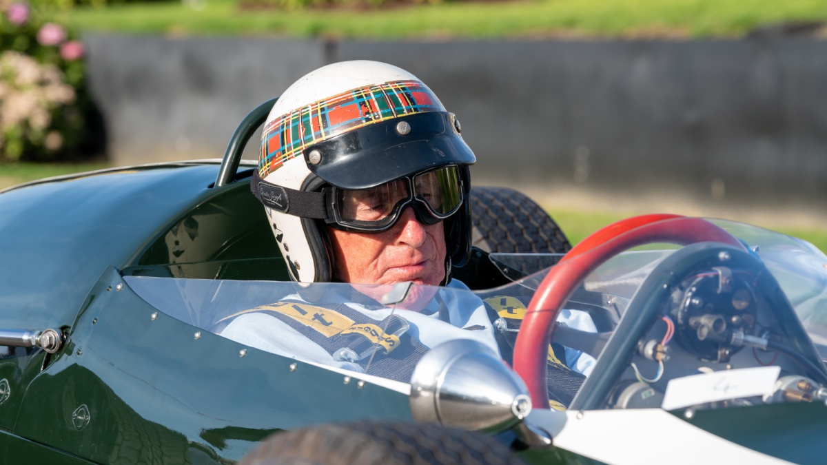 Sir Jackie Stewart, wearing his trademark tartan and white helmet, sits behind the red steering wheel of a classic race car at the annual vintage Revival event.