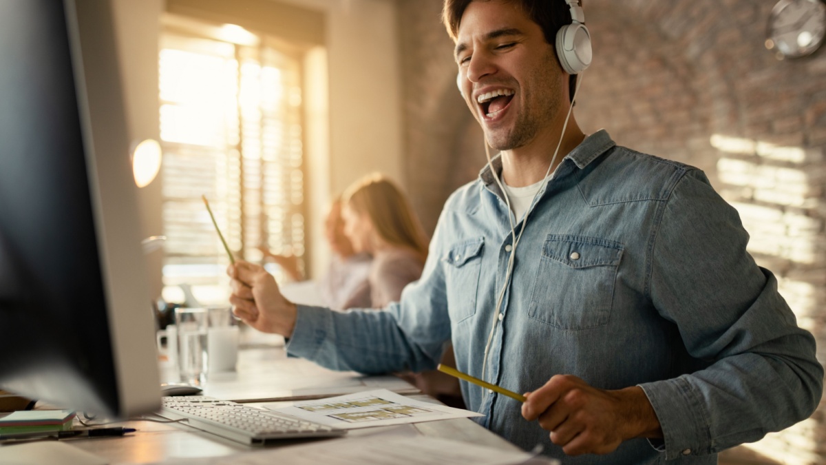 Happy freelance worker having fun while working at office desk and listening music over headphones. His colleagues are in the background.