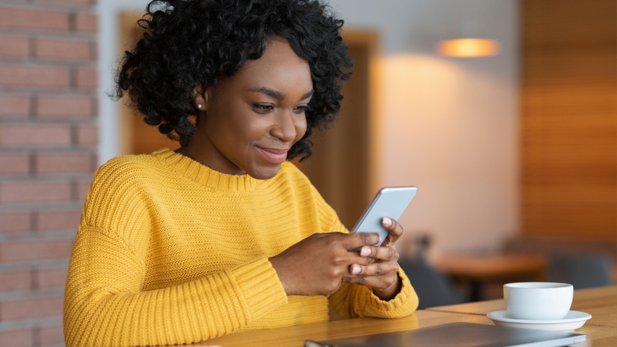 Busy cheerful black girl chatting at cafe, using modern smartphone and drinking tea.