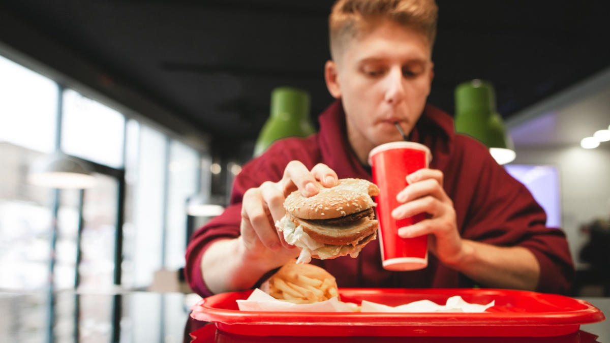 Tray with fast food, french fries, burger in the hands of a young man drinking a drink from a red glass.
