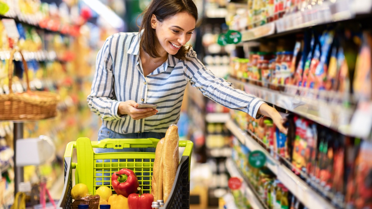 Shopping. Young Smiling Woman Holding And Using Mobile Phone Buying Food Groceries Standing In Supermarket.