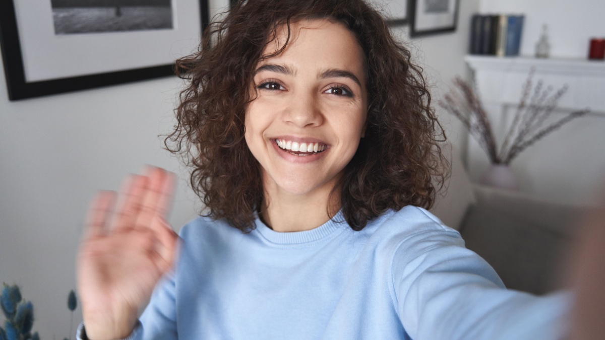 Happy pretty hispanic gen z teen girl vlogger holding smartphone waving hand, talking to camera shooting vlog, making video call at home talking by virtual mobile video call chat app. Webcam view.