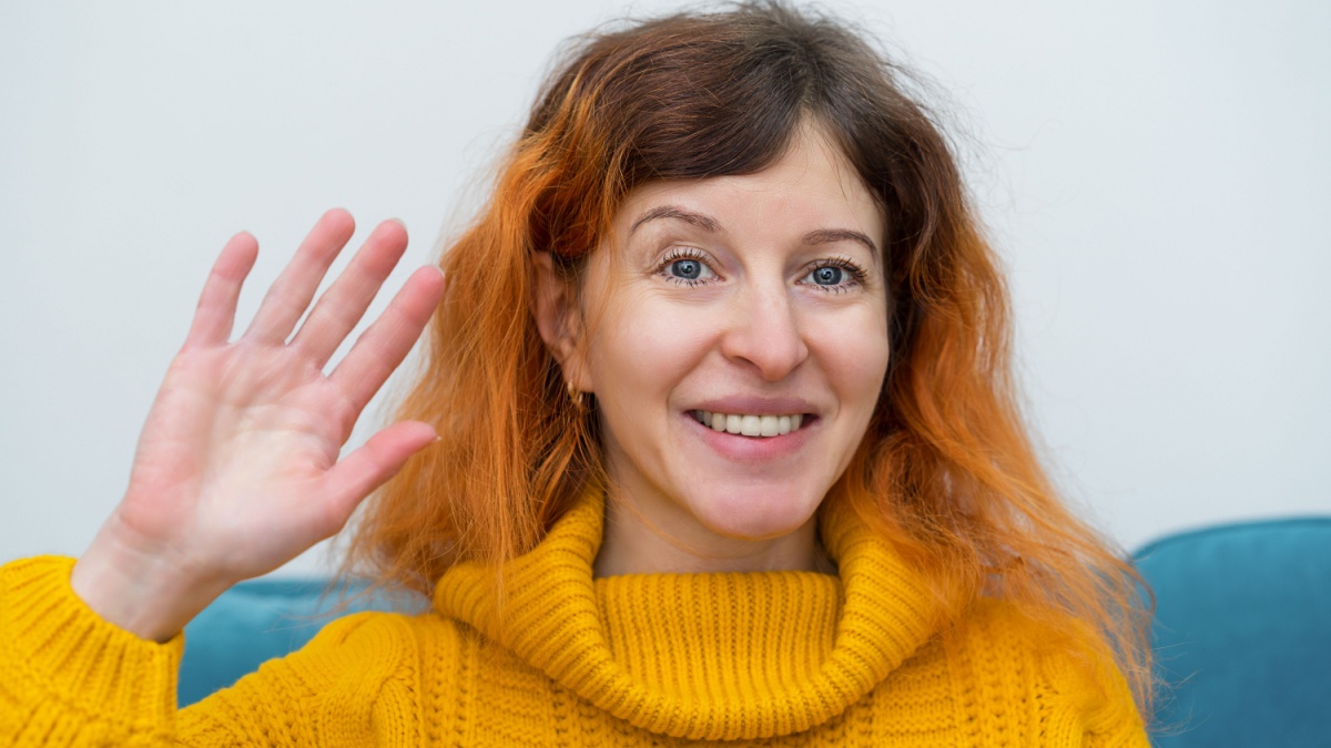 Happy smiling middle-aged woman with red hair wearing a yellow sweater talking to webcam.