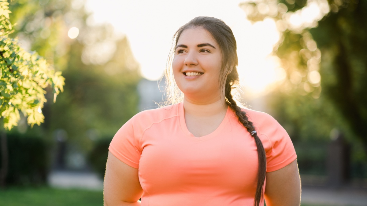 Obesity people. Plus size woman. Body positive. Weight confidence. Joyful chubby young obese overweight girl smiling outdoors in defocused green park street landscape.