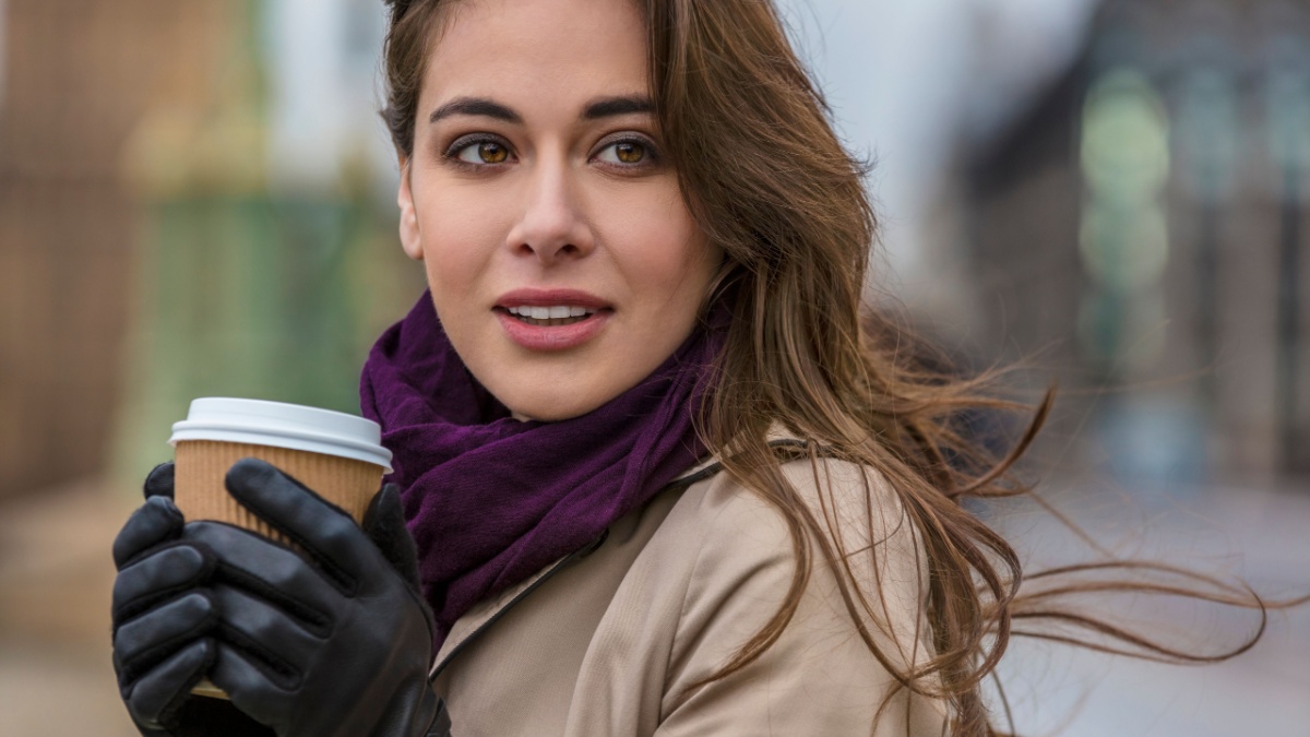 Girl or young woman drinking coffee or tea in a disposable cup outside wearing gloves, a scarf and raincoat in cold wet weather.