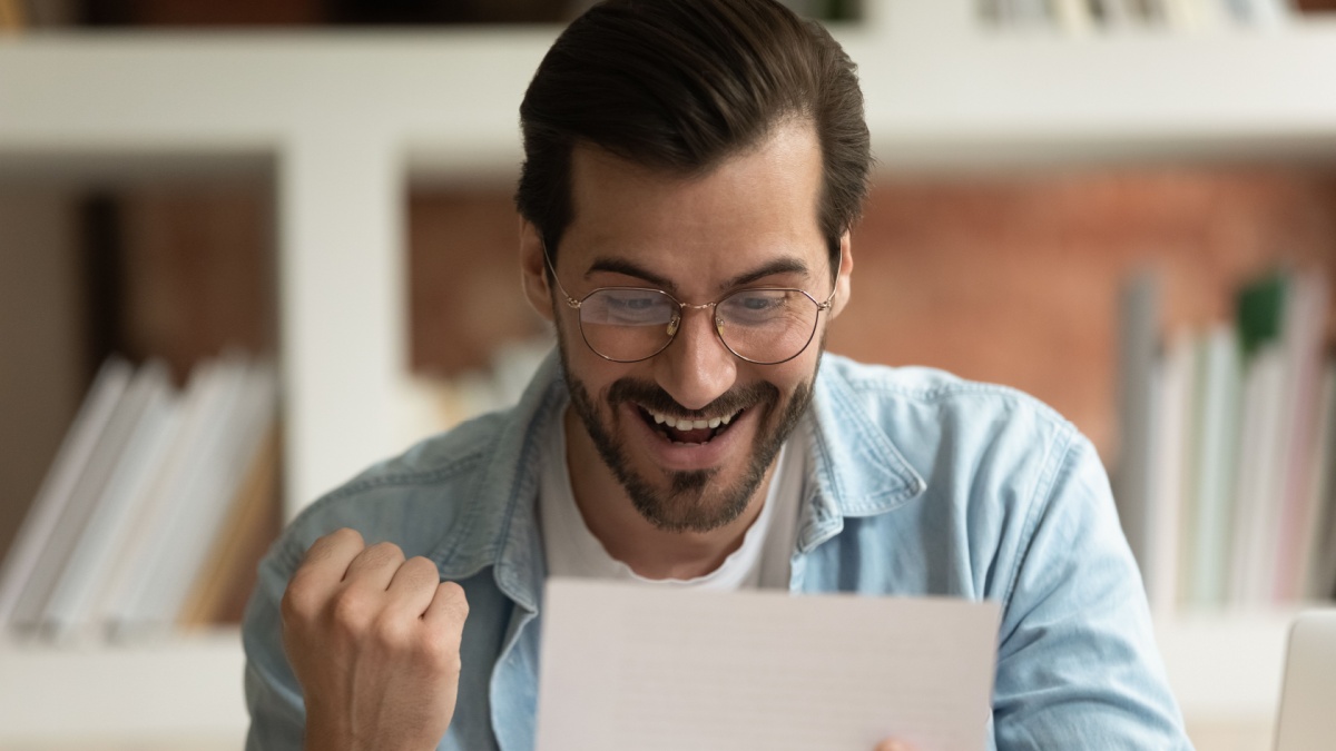 Excited business man sit indoor read postal mail letter feels overjoyed by great news, clench fist makes yes gesture.