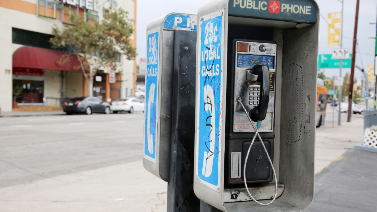 An old, dirty coin operated payphone is shown outside along a city sidewalk during a sunny day. Public Pay Telephone for use by anyone.