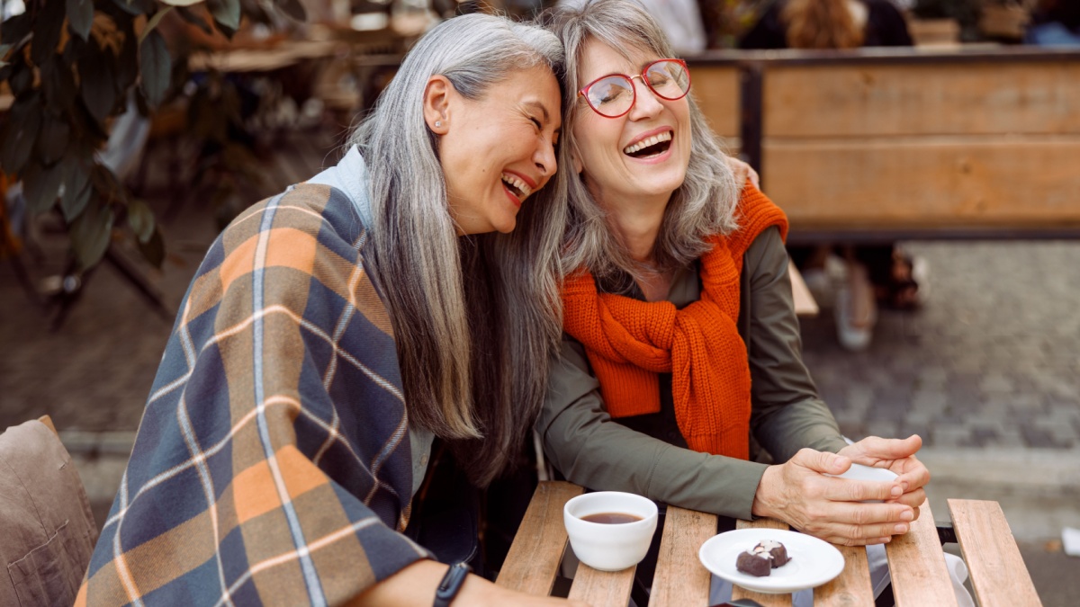 Pretty senior Asian woman puts head on laughing friend shoulder resting together at table in street cafe on nice autumn day.