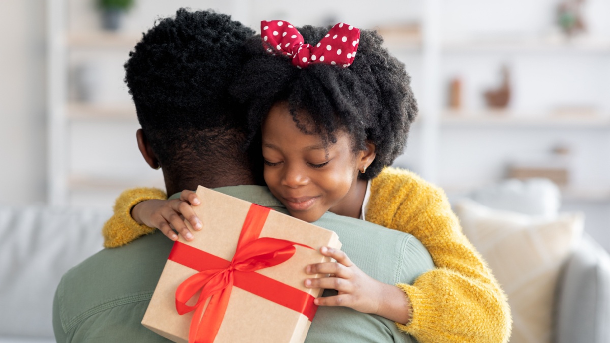 Smiling little black girl holding gift box and embracing dad, loving daughter greeting daddy with Father's day or birthday, happy african american family bonding together at home, closeup shot.