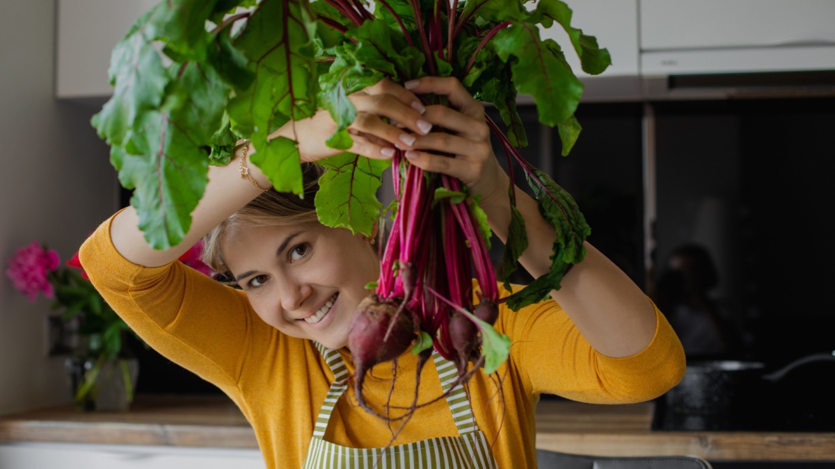 Blond smiling woman holding bunch of beetroots with leaves, cooking fresh organic salad in the kitchen on summer weekend.