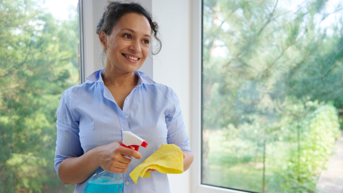 Beautiful multi-ethnic woman housewife, homemaker, maid wearing blue casual shirt, spraying detergent on a yellow cloth while washing panoramic windows in veranda. Spring cleaning and housekeeping.