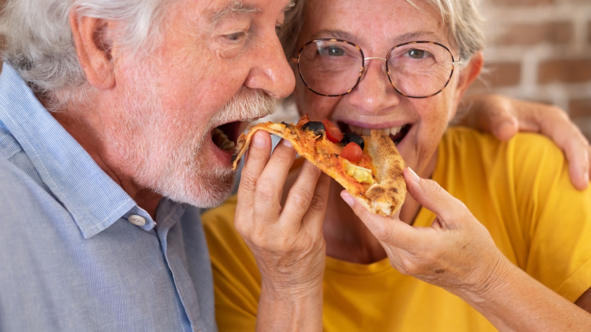 Senior couple having fun together eating pizza at home, sharing the same slice, brick wall background.