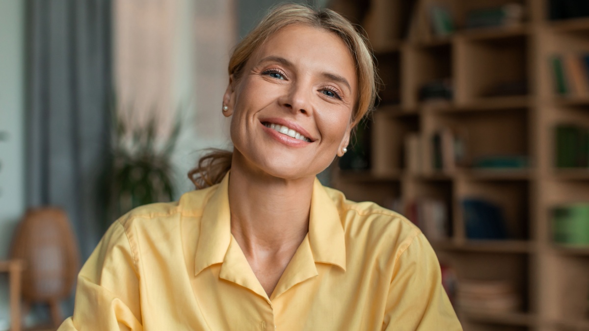 Portrait of positive middle aged female psychologist talking to camera and smiling during therapy session in office. Friendly psychotherapist posing in cabinet.