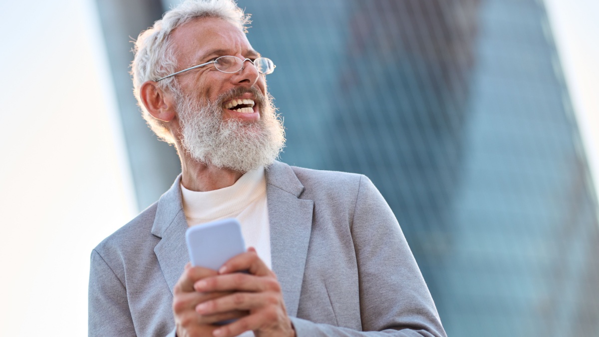 Happy older mature stylish professional business man, smiling senior 60 years old businessman wearing suit holding smartphone using mobile cell phone tech standing outdoor in big city office district.