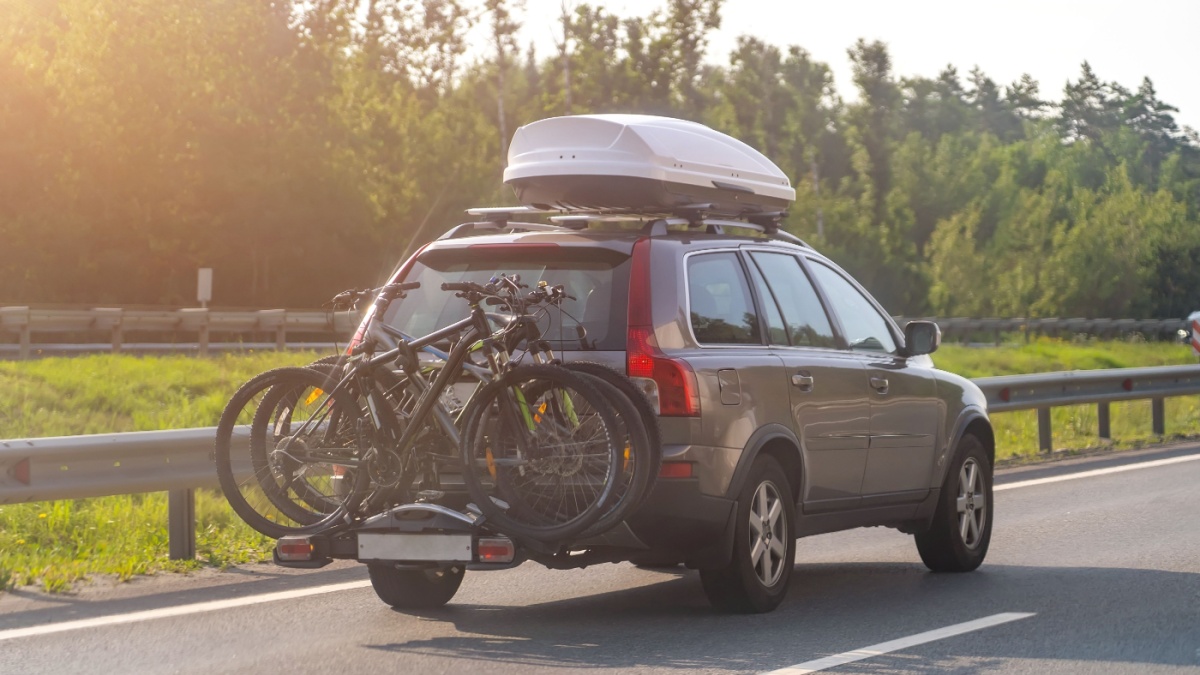 Bikes fastened on bicycle holder mounted on back side of car on country road. Brown car with roof luggage box and trunk bike rack driving on highway.