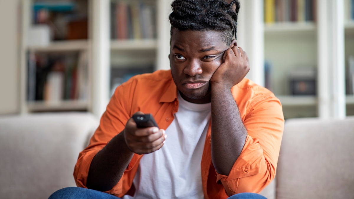 bored chubby young african american guy with braids sitting on couch in living room, switching channels on tv, touching his face, looking for something.