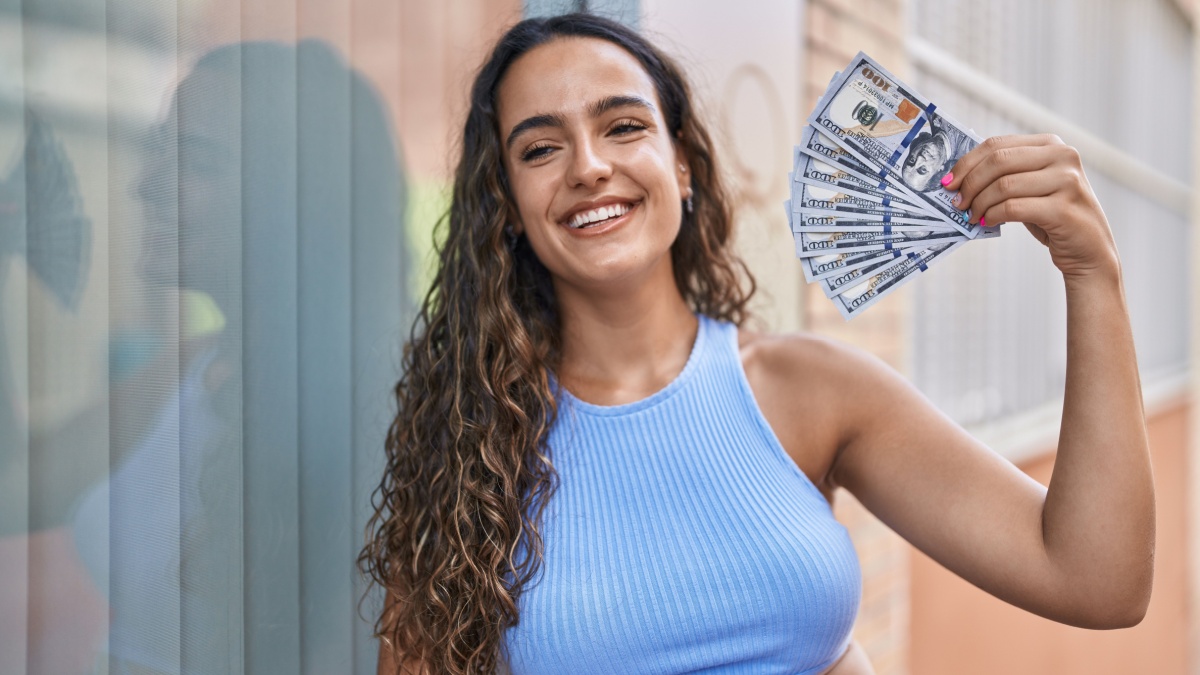 Young beautiful hispanic woman smiling confident using dollars as a handfan at street.