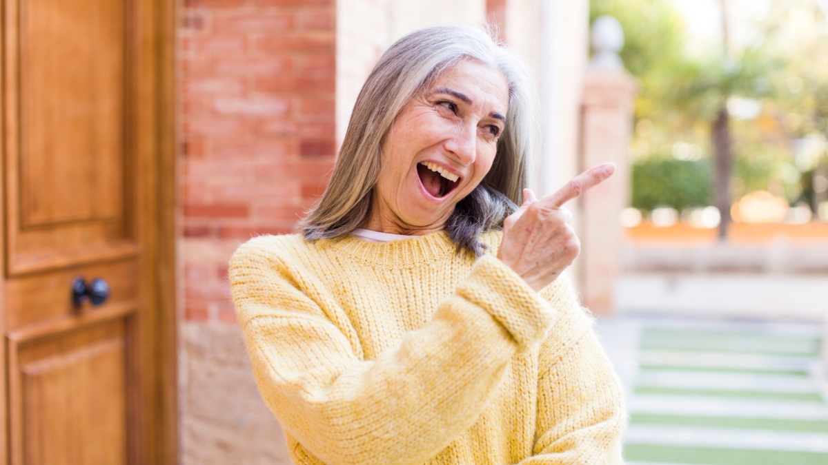 senior retired pretty white hair woman feeling joyful and surprised, smiling with a shocked expression and pointing to the side.