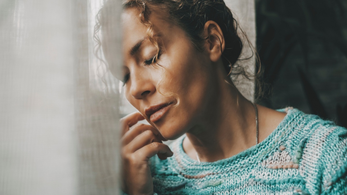 Exhausted and sad woman alone at home near the window with closed eyes and depressed expression on face.