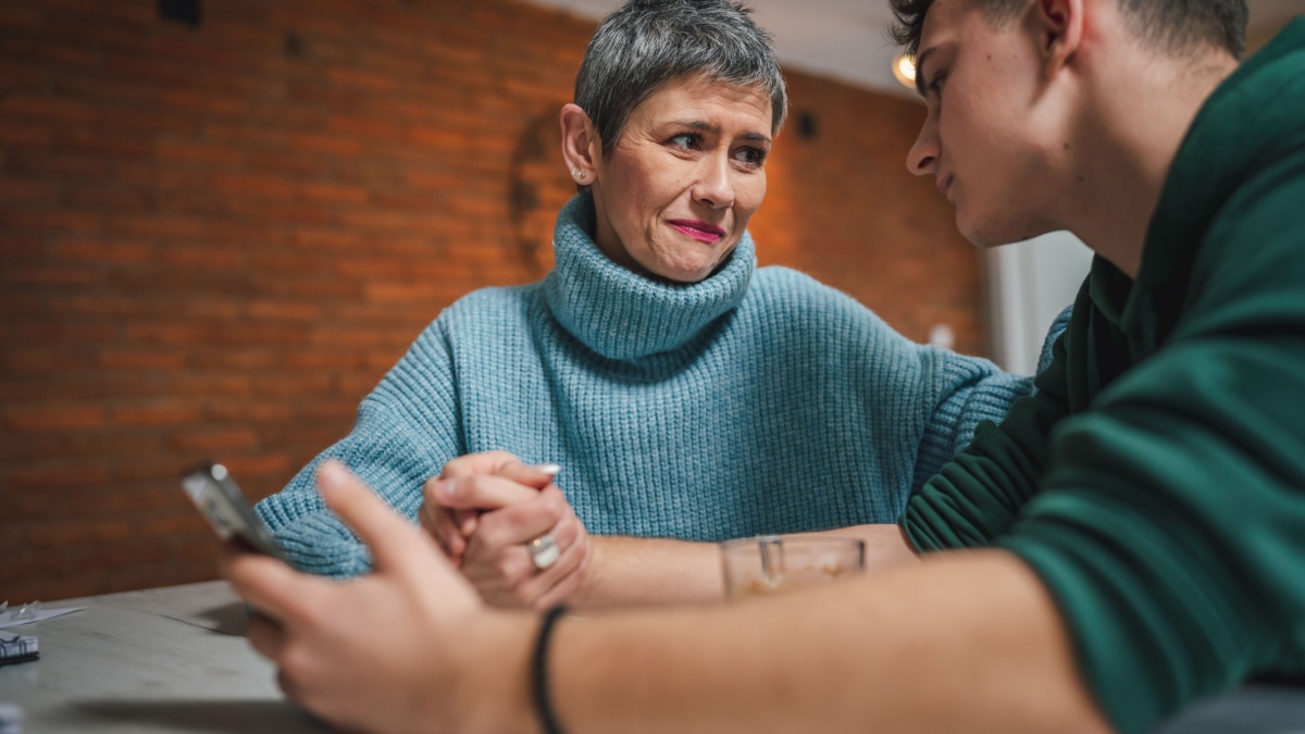teenage boy and mature caucasian woman sit together at the kitchen at home talk mother and son or relatives support solving problem share experience and opinion boy hold mobile phone smartphone.