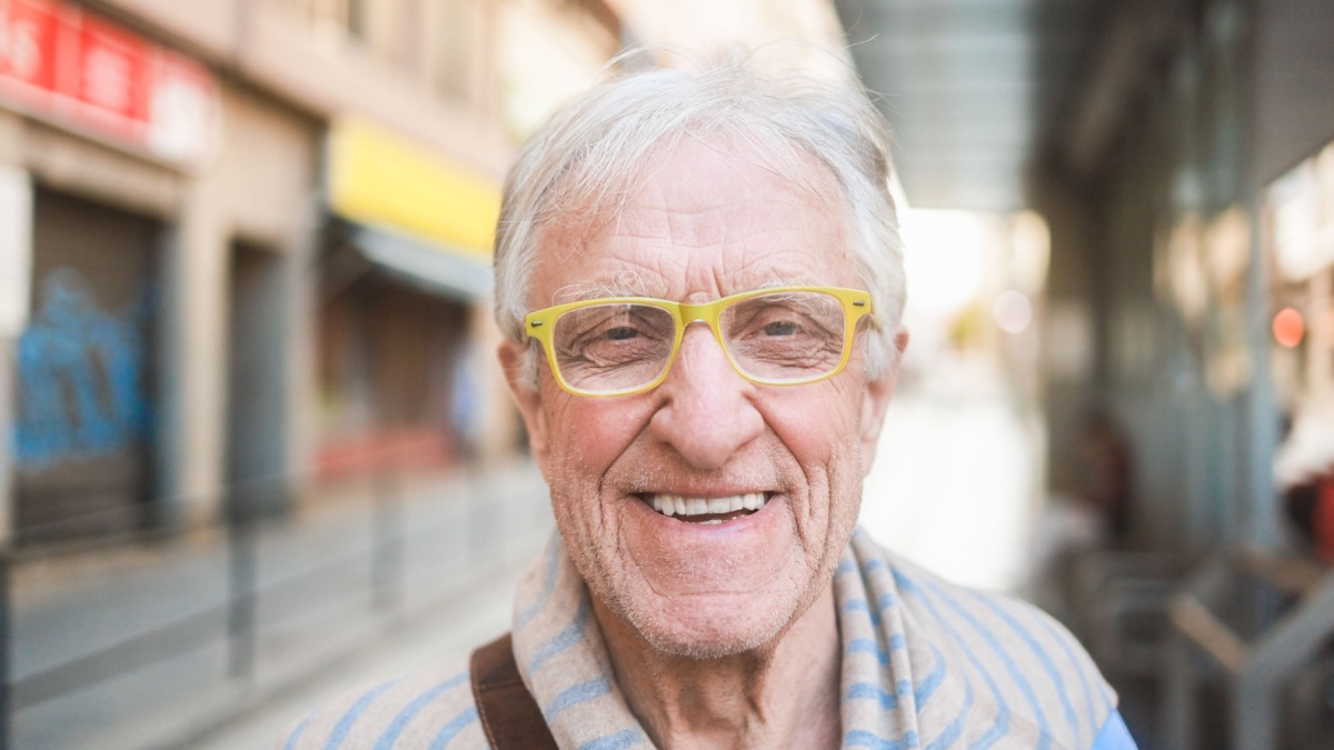 Senior man smiling in front of camera while waiting tram at city station - Citizen lifestyle and transportation concept - Focus on mouth.