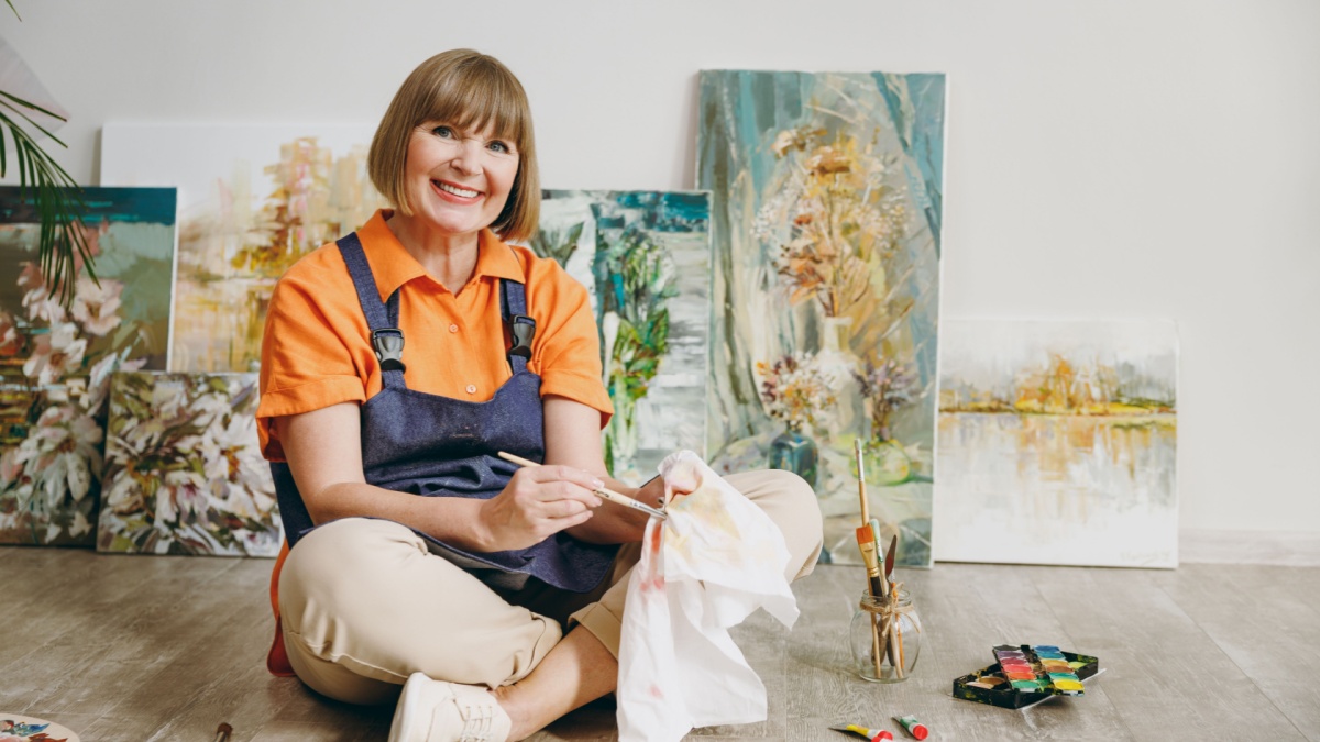 Full body elderly fun artist woman 50 years old wears casual clothes sit on floor painting on canvas artwork wiping cleaning brushes spend free spare time