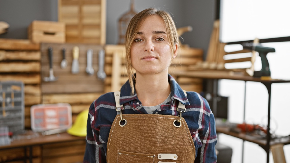 Young, serious blonde woman carpenter stands tall in carpentry studio, bringing beauty and professionalism to woodworking industry.