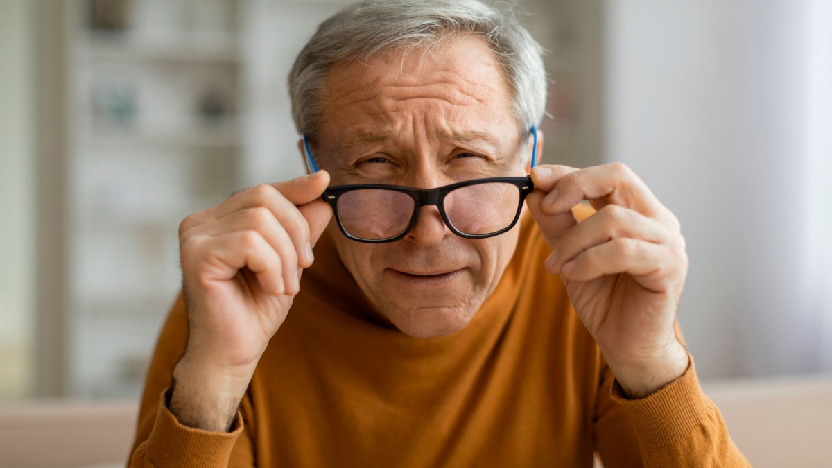 Tired grey-haired elderly man removing eyeglasses and squinting, sitting on couch, home interior, closeup, copy space. Vision care for senior people.