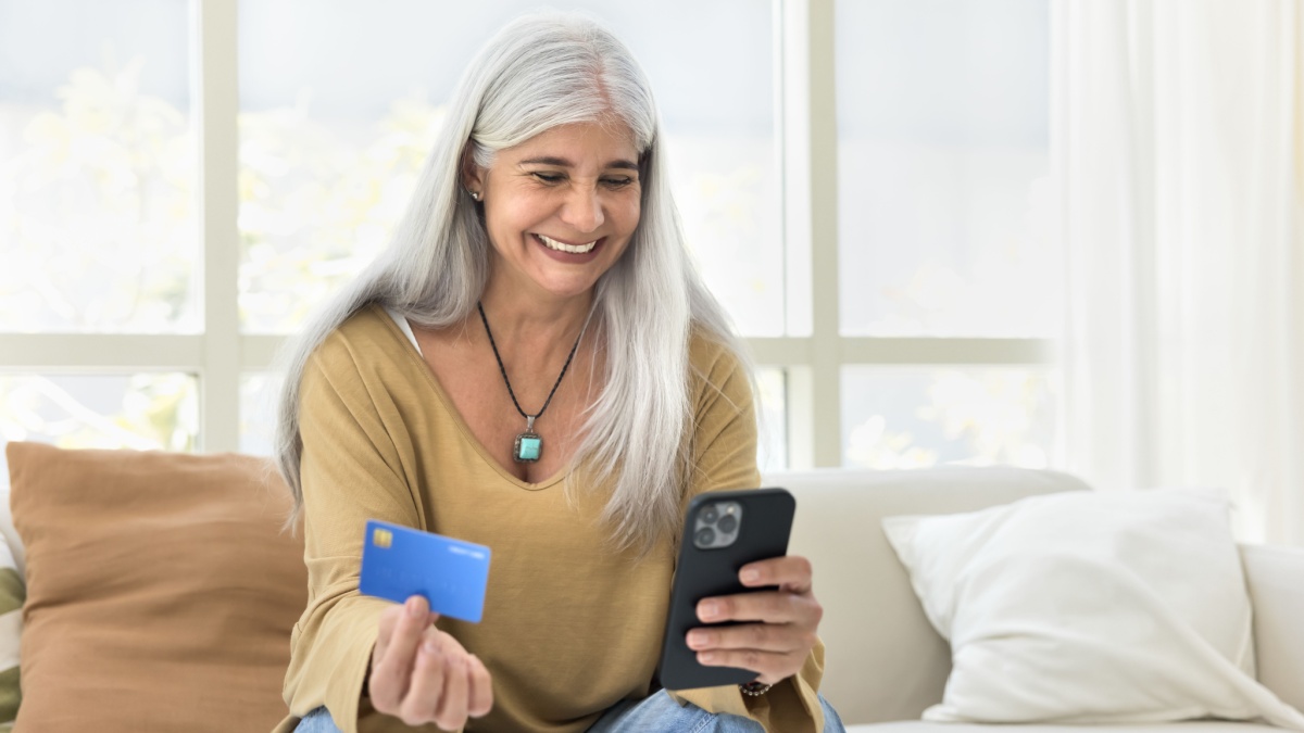 Cheerful grey haired senior Latin woman making money transaction with online financial service for payment, holding mobile phone, paying for Internet.