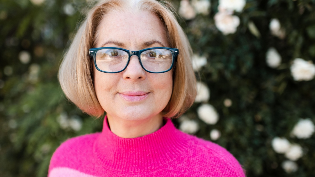Smiling blonde mature woman 50-60 year old with gray hair wearing vision glasses and pink knitted sweater over blooming rose flowers outdoor.