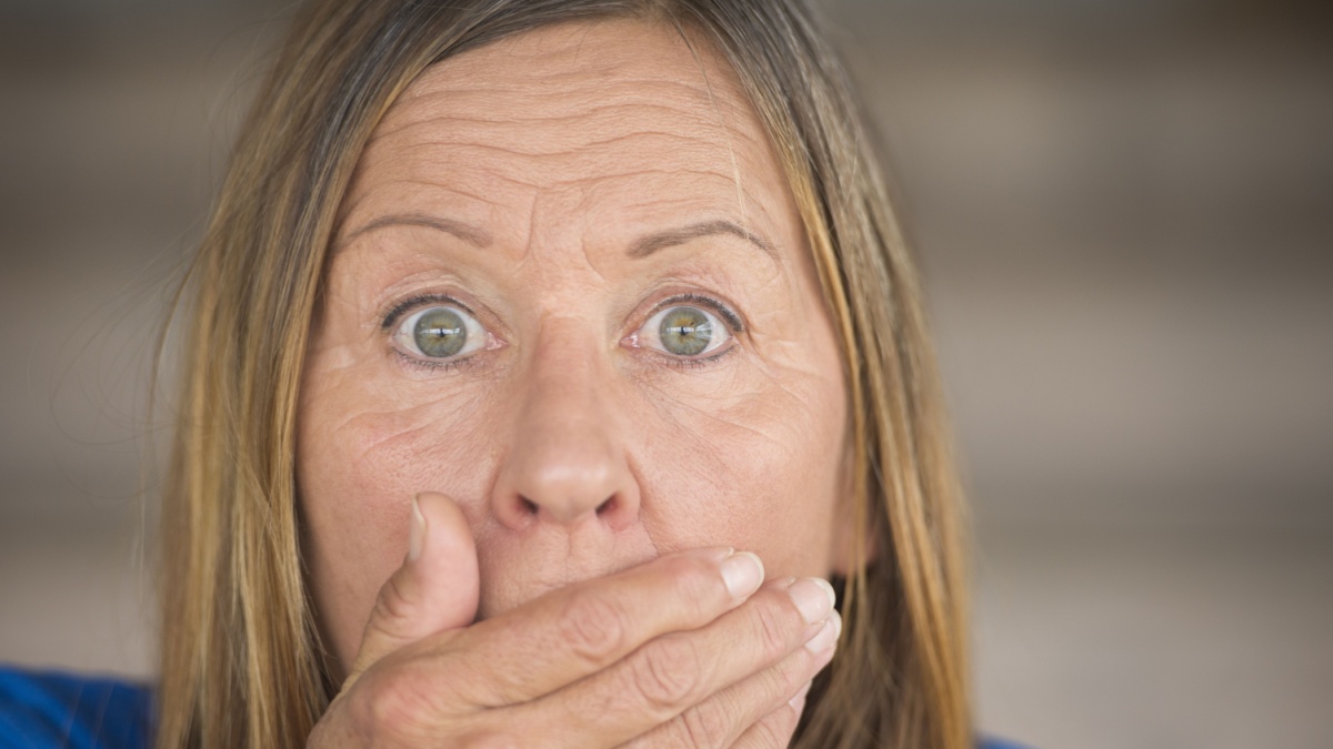Portrait attractive mature woman with shocked, surprised, anxious facial expression, covering mouth with hand, blurred background.