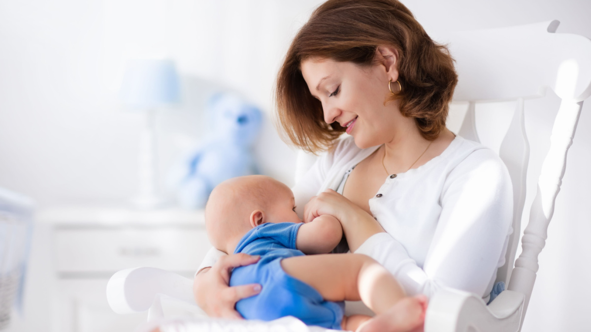 Young mother holding her newborn child. Mom nursing baby. Woman and new born boy in white bedroom with rocking chair and blue crib. Nursery interior. Mother playing with laughing kid. Family at home