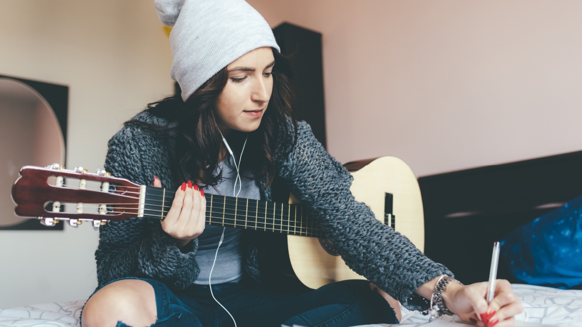 Young beautiful eastern woman sitting on her bed in the bedroom holding guitar composing a song - musician, songwriter, composer concept