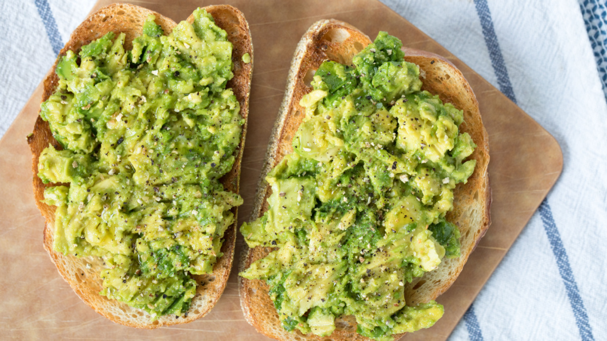 Over head avocado toast on slices of toasted French loaf bread topped with ground lemon pepper and sprinkles of kosher salt on wood cutting board on top blue and white striped linen cloth