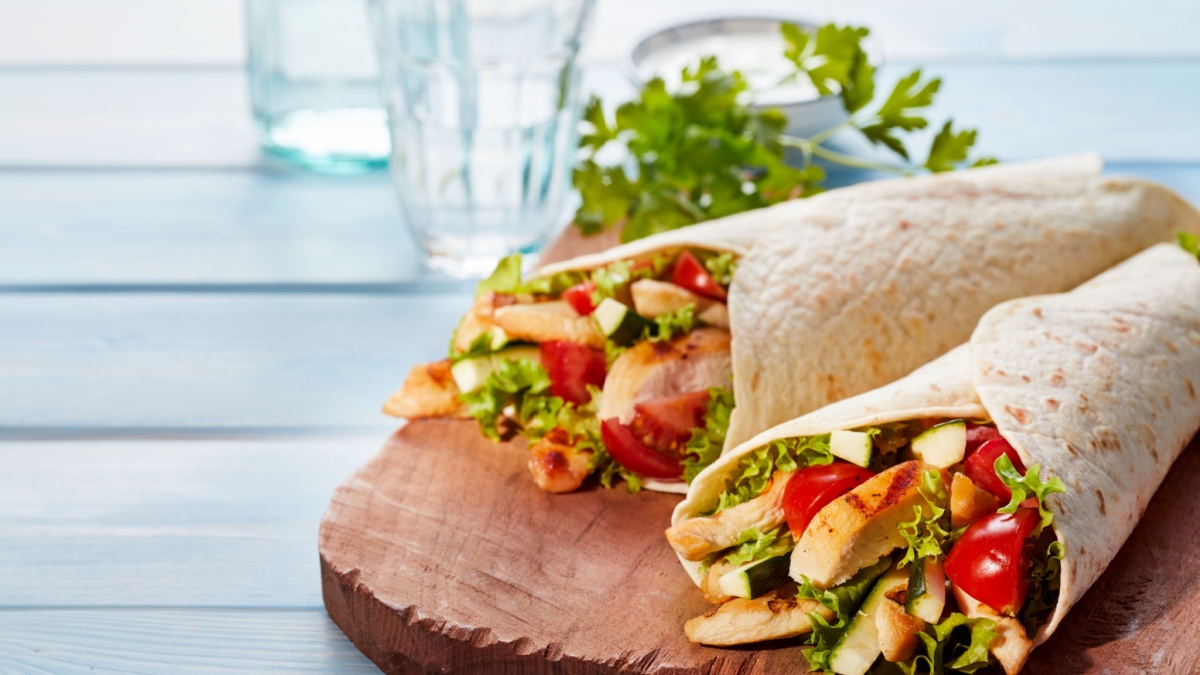 Two fresh chicken and salad tortilla wraps on wooden cutting board with glasses in background
