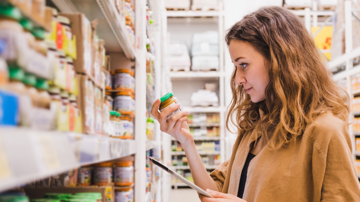 9 Essentials Frugal People Proudly Exclude From Their Shopping List 3 Young beautiful woman with a tablet picks baby food in a supermarket, the girl is studying the composition of the product close-up