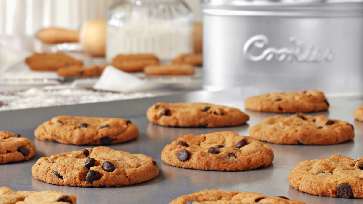 Baking still life of freshly baked chocolate chips cookies on non-stick cookie sheet with canisters, cooling rack and baking supplies in background. Closeup with shallow dof.