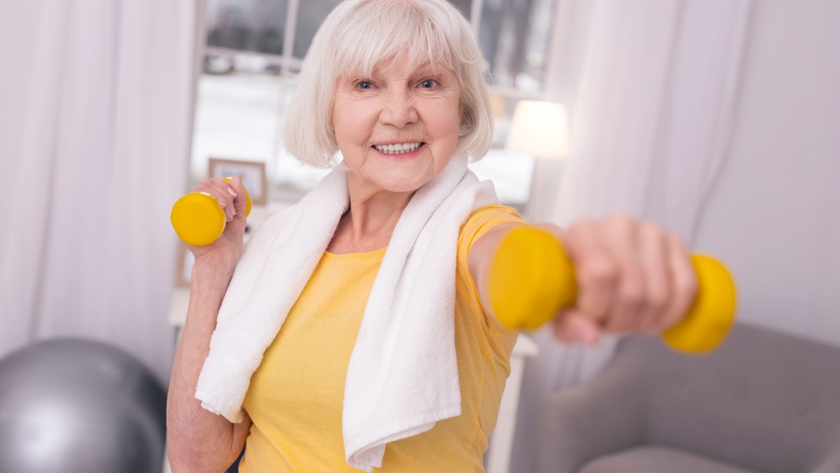 Gaining strength. Upbeat elderly woman with a towel around her neck exercising with a pair of dumbbells and smiling.