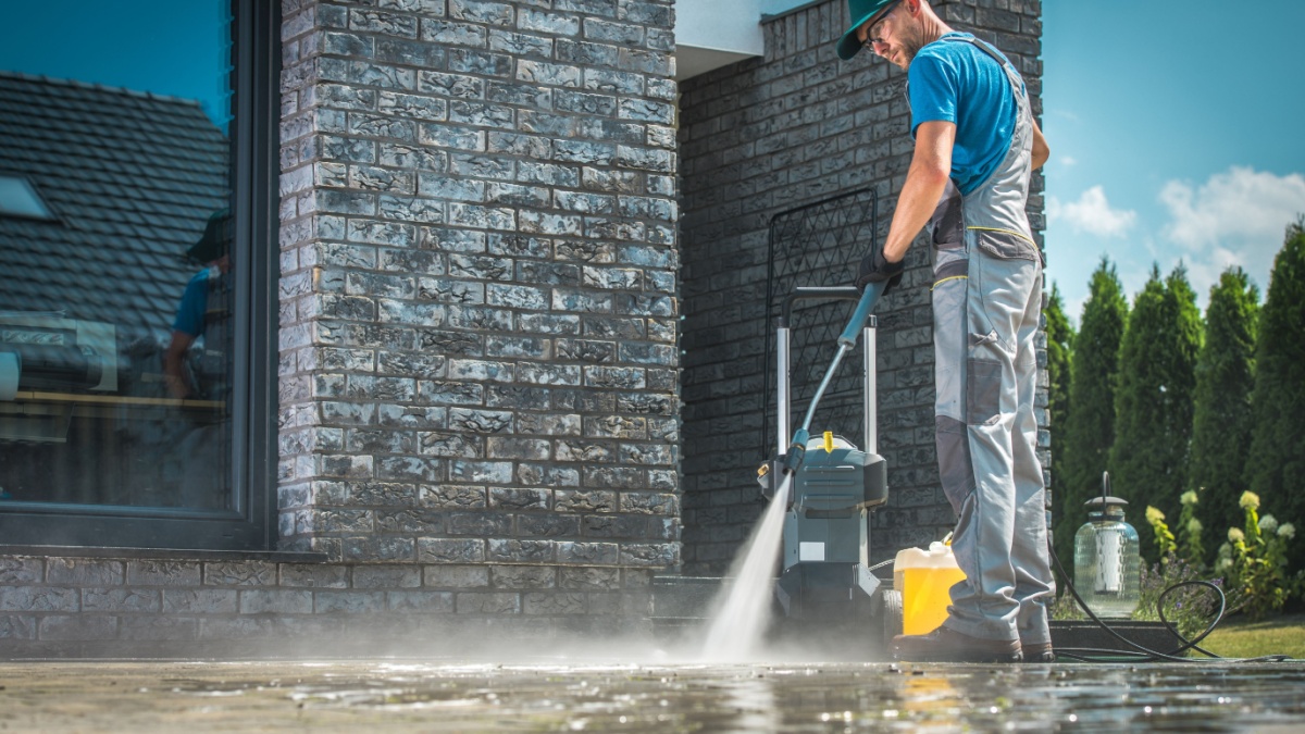 Pressure Washer Cleaning in Front of the House. Caucasian Men in His 30s Washing Concrete Bricks Driveway in Sunny Summer Day. Cleaning Around the House Concept
