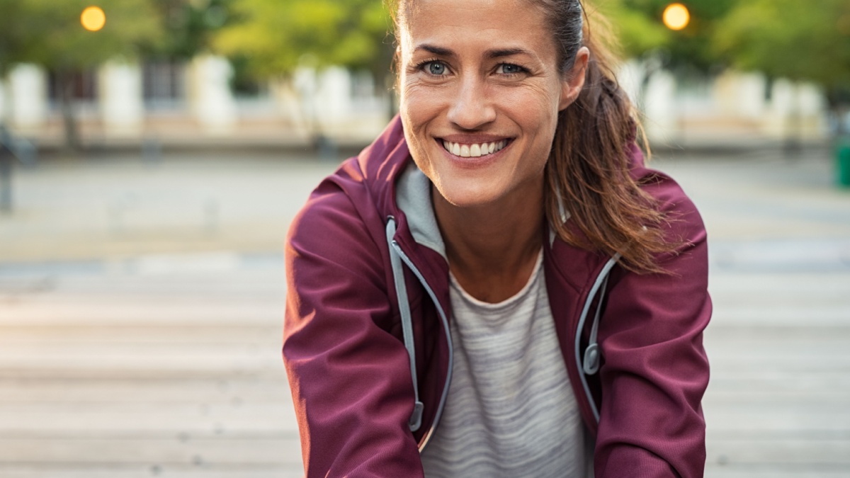 Portrait of smiling woman sitting on floor of city street after running. Healthy mature runner resting after workout exercise and looking at camera. Active sporty woman enjoying outdoors in autumn.