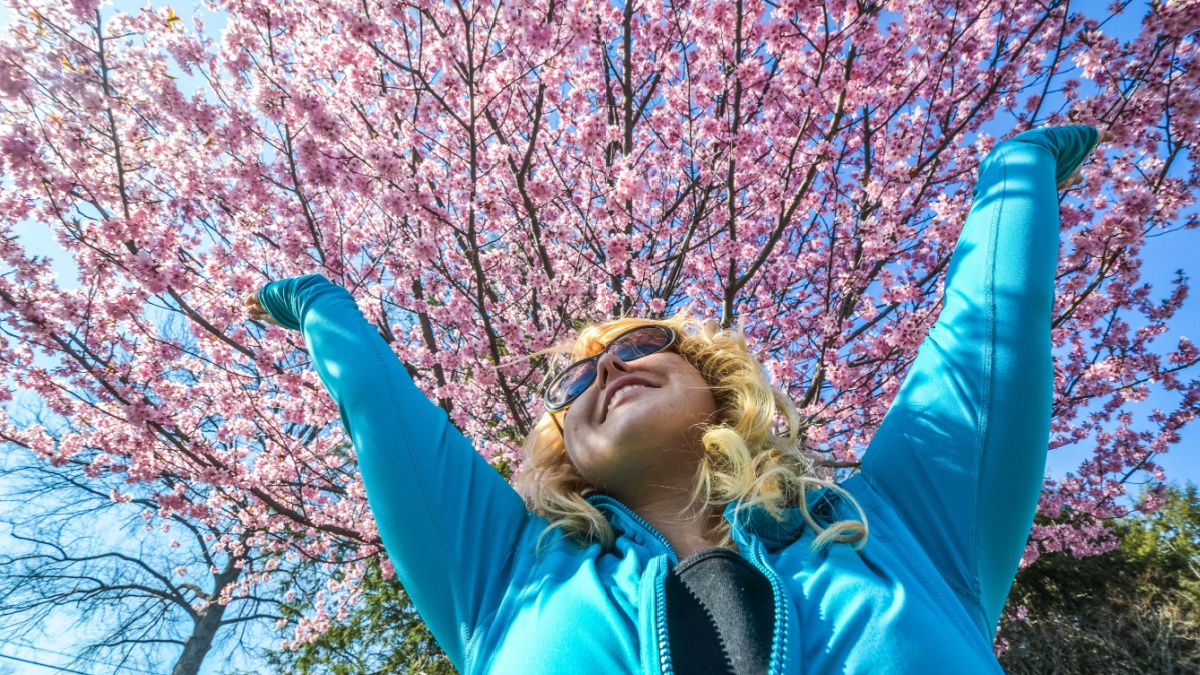 Blonde woman raises her arms up next to a beautiful pink cherry blossom tree and smiles.