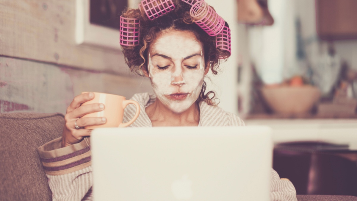 Rare and funny portrait of beautiful woman with facial mask and curlers hair taking a coffee at home while use internet with modern laptop computer - home.
