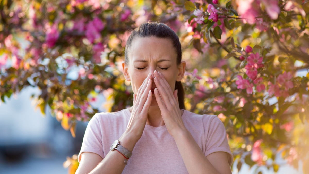 Young pretty woman sneezing in front of blooming tree. Spring allergy concept.