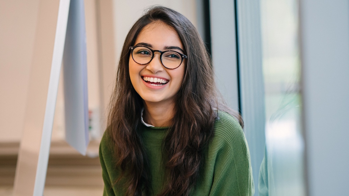 A close up head shot portrait of a preppy, young, beautiful, confident and attractive Indian Asian woman in a green sweater and spectacles in a classroom or office. She is smiling happily.
