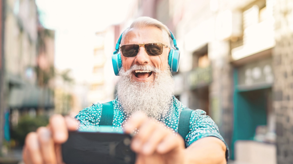 Senior bearded man taking selfie with mobile phone while listening favorite playlist with headphones.